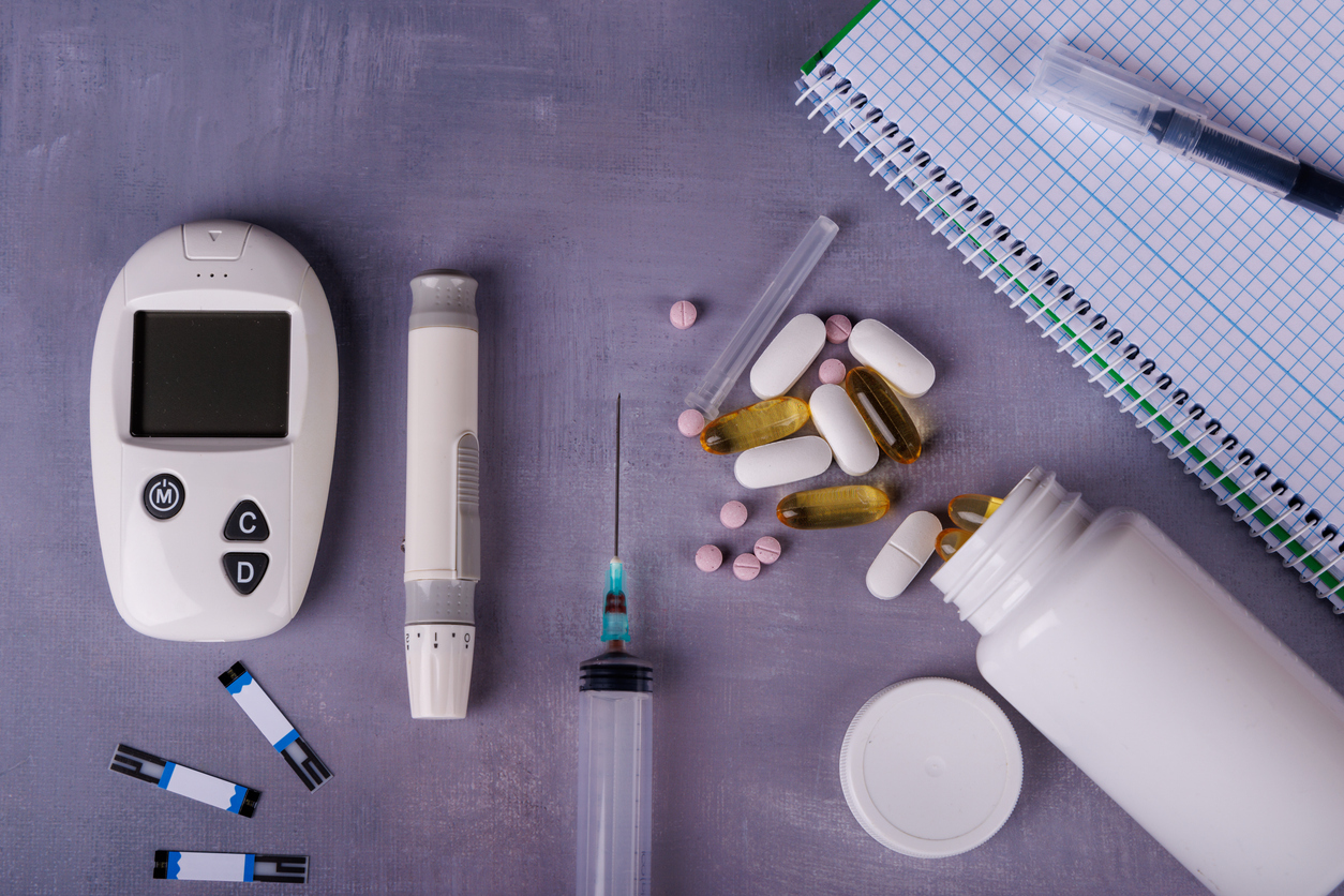 A top-down view of medical supplies: a glucometer, thermometer, syringe, white pills, and a notebook neatly organized on a concrete table